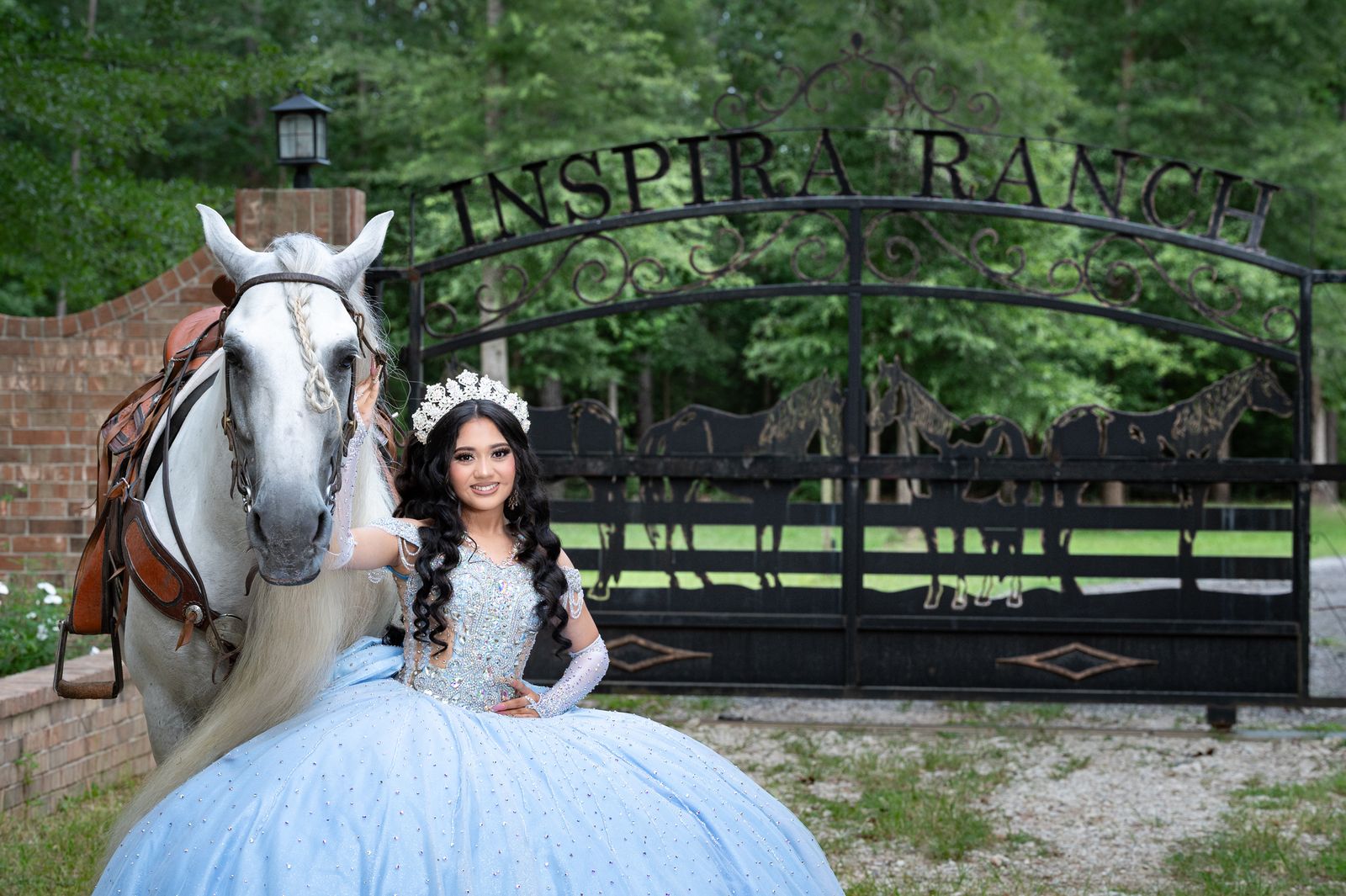 Quinceañera in a blue gown beside a saddled white horse at the Inspira Ranch gate