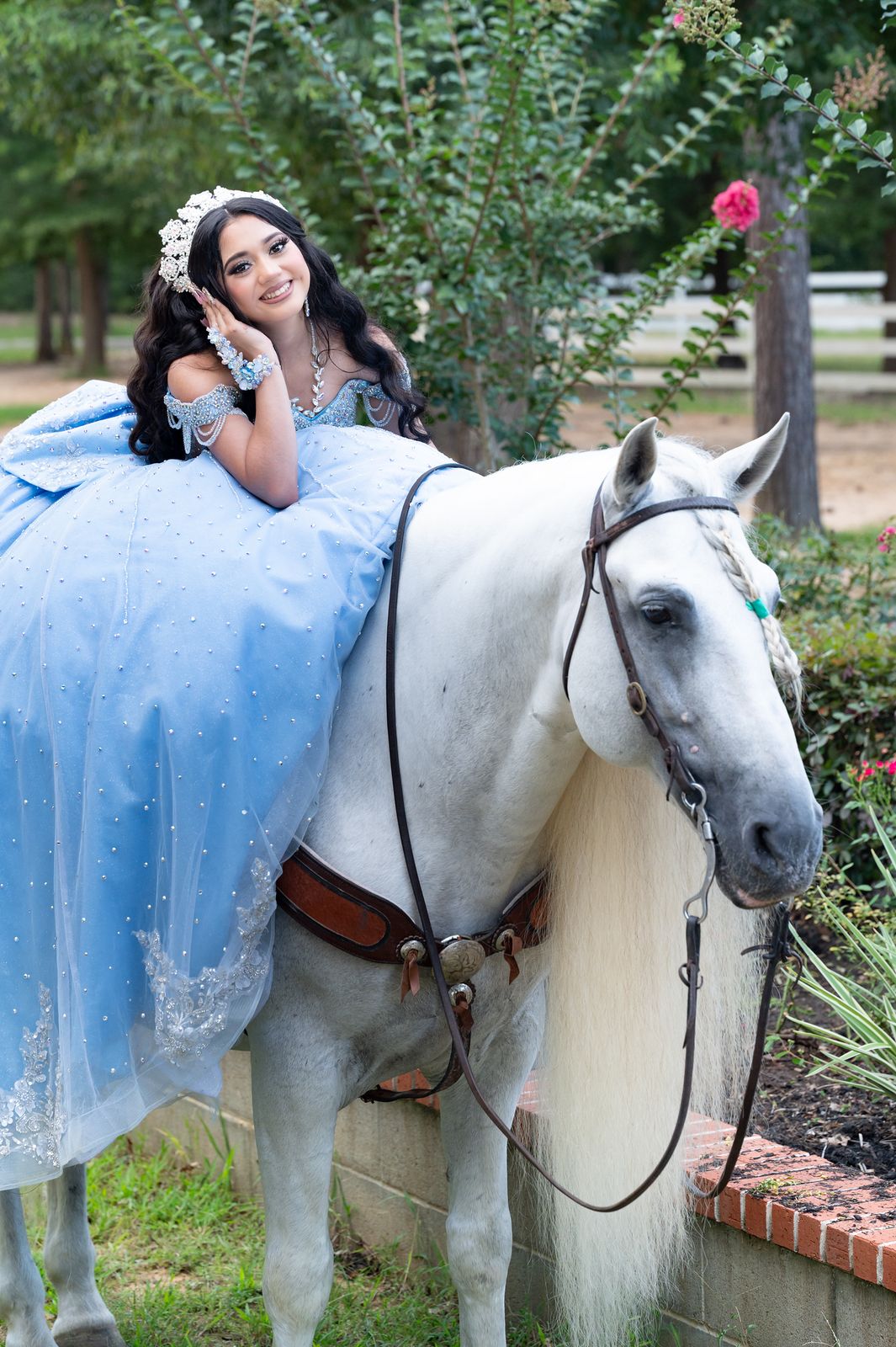 Quinceañera resting on a white horse in a flowing blue gown by the garden wall