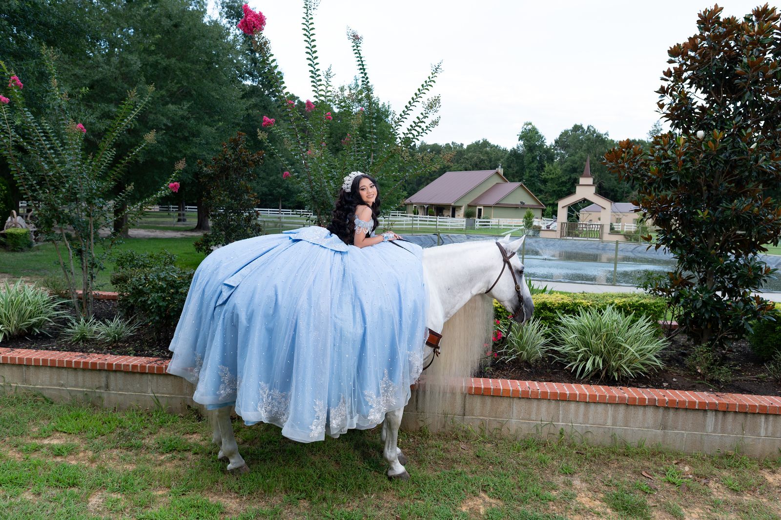 Quinceañera resting on a white horse with the pond and ranch houses behind